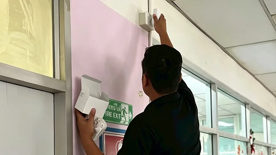 Technician mounting an MK107 BLE plug gateway on a pink hospital corridor wall near a fire exit sign